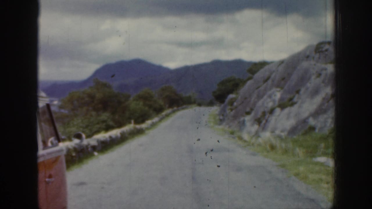 1961 Ireland home-movie frame from a moving vehicle showing a narrow road, stone wall, rocky slope, and distant mountains under cloud.