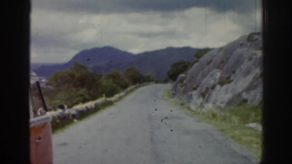 1961 Ireland home-movie frame from a moving vehicle showing a narrow road, stone wall, rocky slope, and distant mountains under cloud.