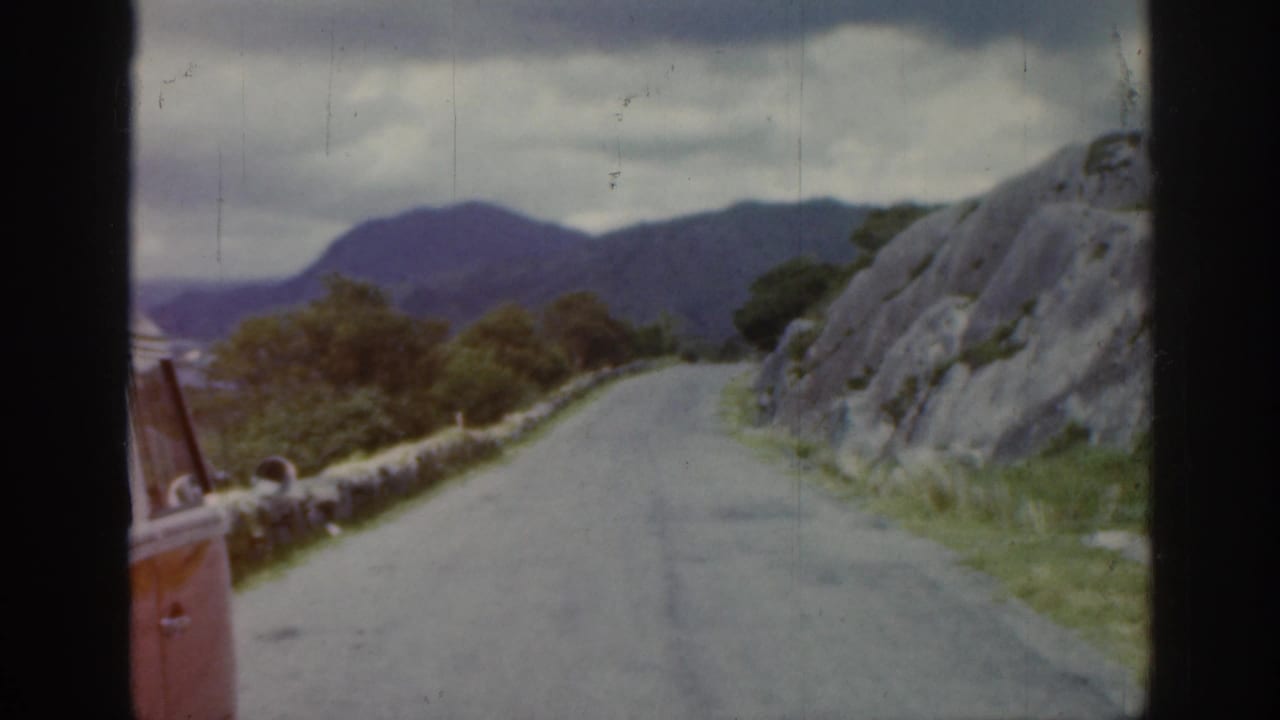 1961 Ireland travel footage frame with a curving road, stone wall, rocky embankment, low vegetation, and hazy mountains beyond.