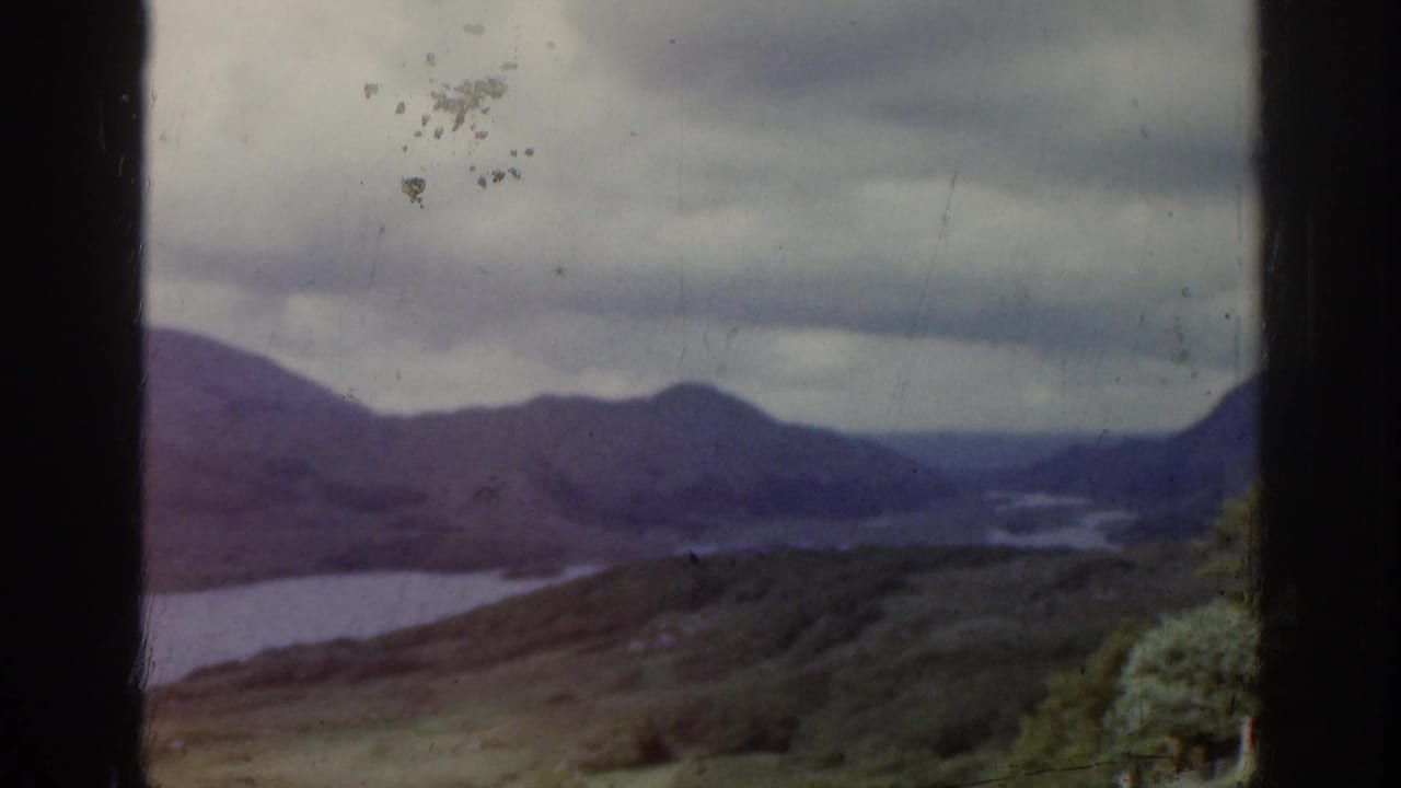 1961 Ireland home-movie view over a lake in a valley, with rolling hills, dark cloud cover, and foreground vegetation.