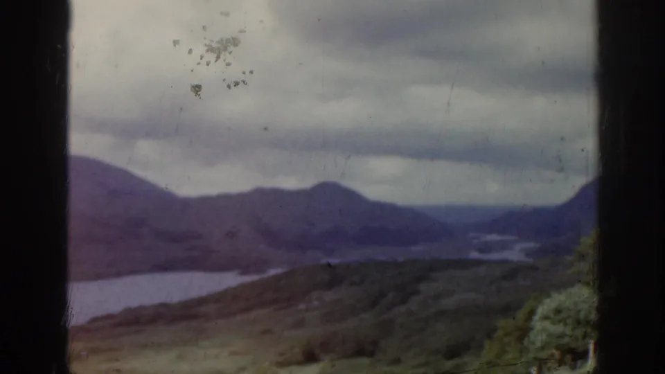 1961 Ireland home-movie view over a lake in a valley, with rolling hills, dark cloud cover, and foreground vegetation.