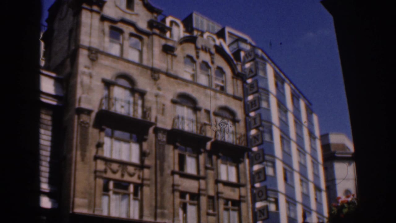 1961 Ireland home-movie frame showing an ornate stone facade, balconies, blue sky, and a modern building with a WASHINGTON sign.