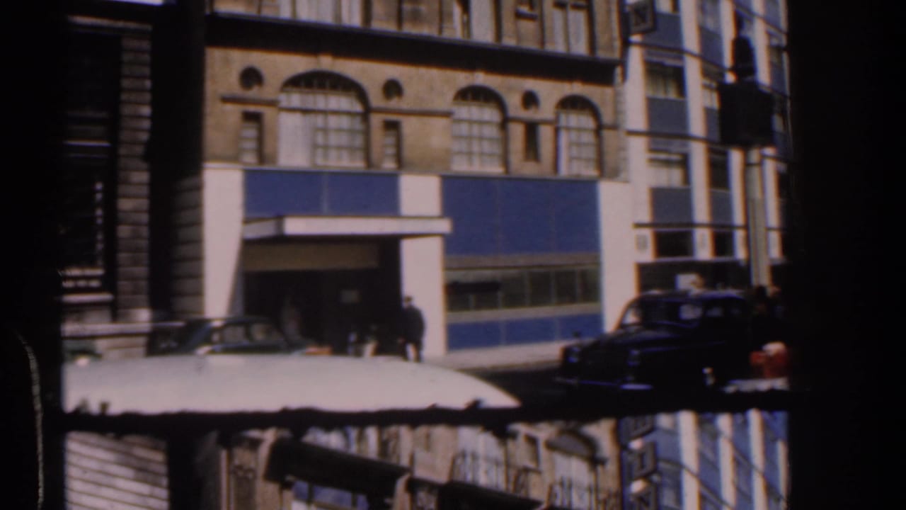1960s London street view with pedestrians near a building entrance, classic cars, and a dark vertical obstruction crossing the frame.