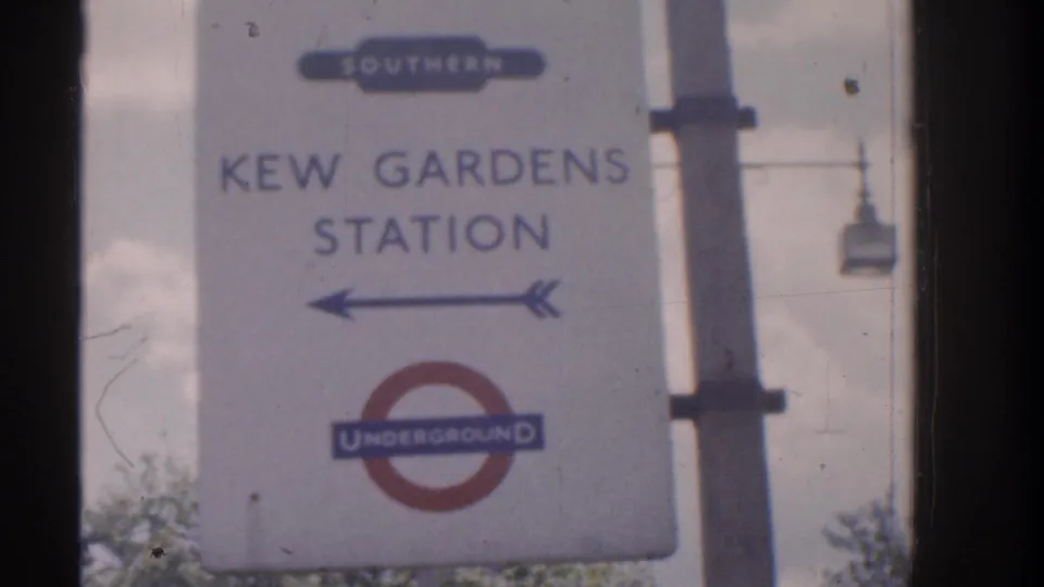 Kew Gardens Station sign showing an Underground roundel, overground wording, and a left-pointing arrow on a pale signboard.