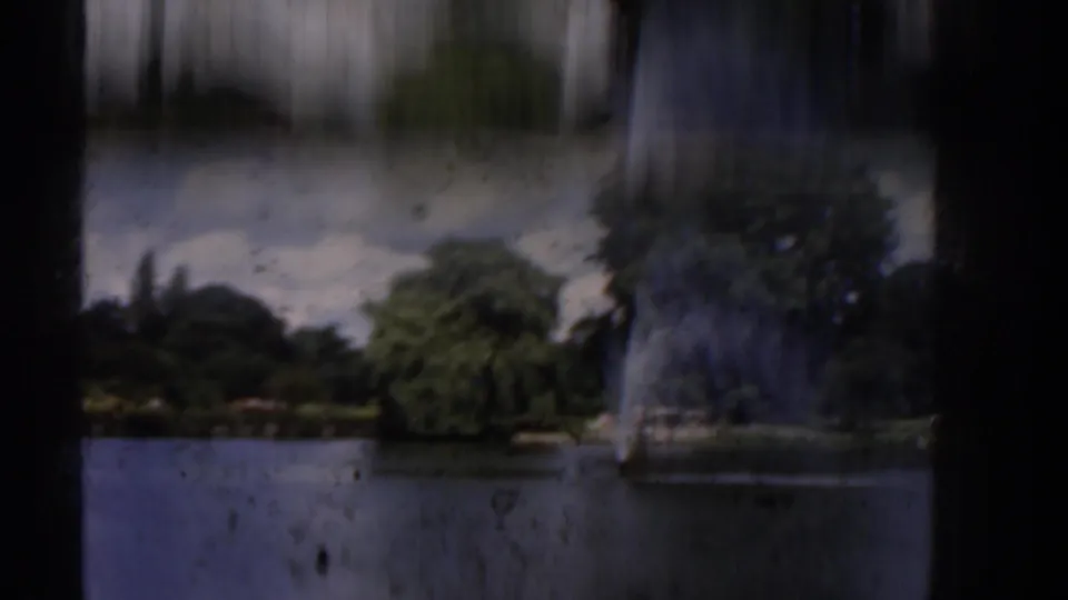 Soft 8mm park view with a lake, fountain spray, trees, and bright sky, partly blurred by camera or vehicle movement.
