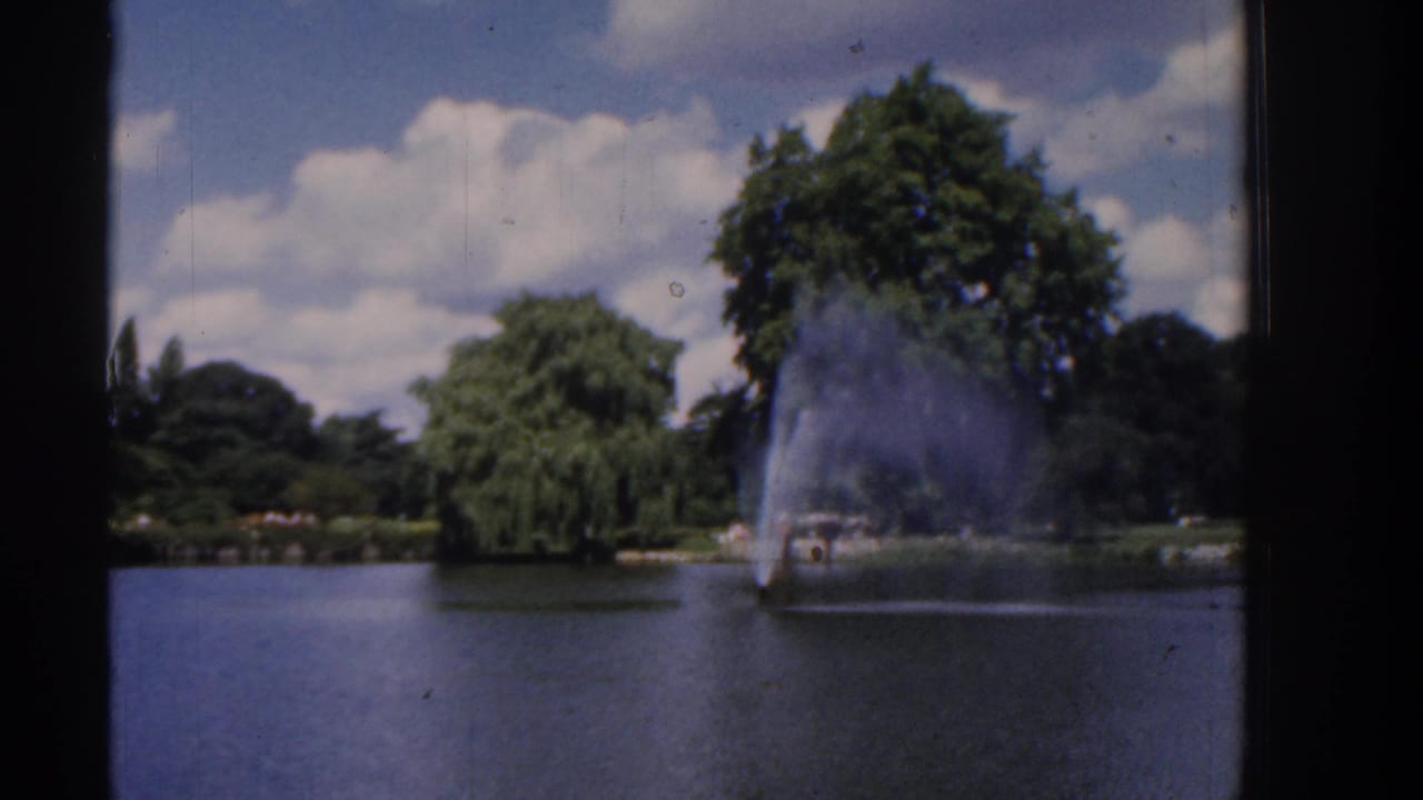 8mm frame of a landscaped lake with a prominent fountain, green trees, and scattered clouds under a blue sky.