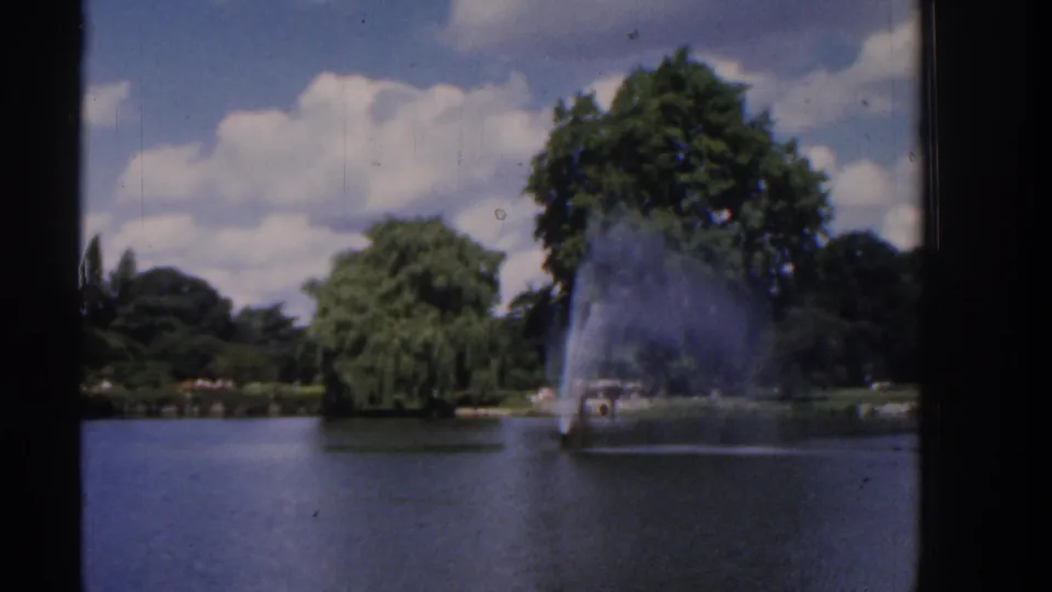 8mm frame of a landscaped lake with a prominent fountain, green trees, and scattered clouds under a blue sky.
