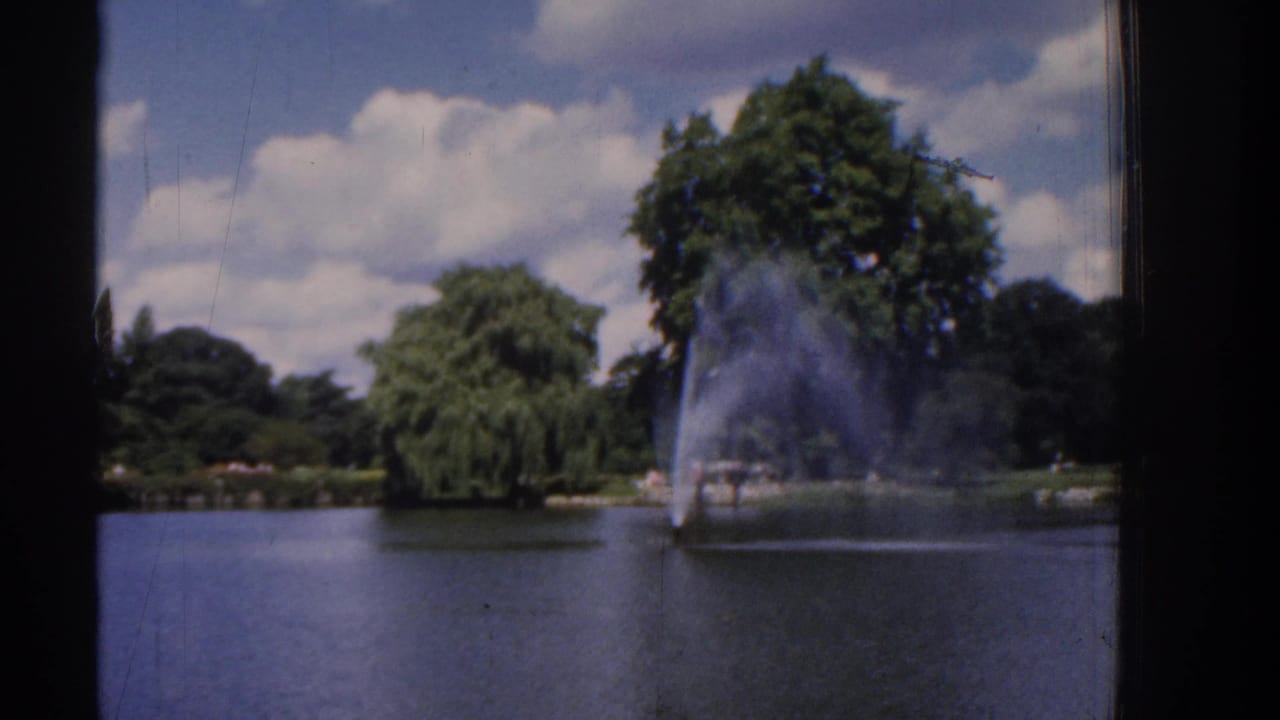 1961 Ireland home-movie frame showing a park pond, distant fountain, leafy trees, clouds, and dark film-frame edges.