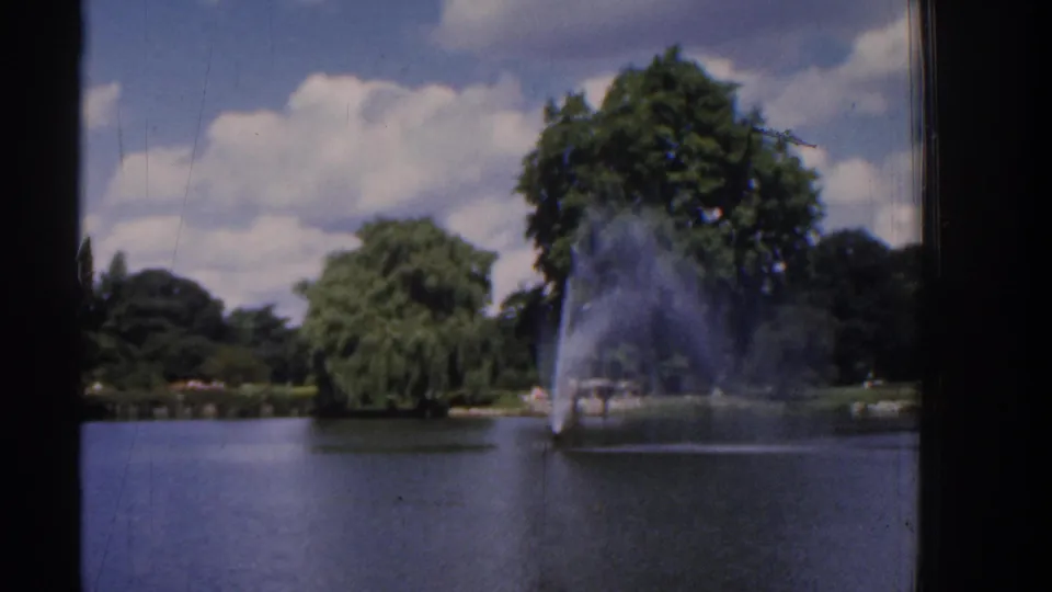 1961 Ireland home-movie frame showing a park pond, distant fountain, leafy trees, clouds, and dark film-frame edges.