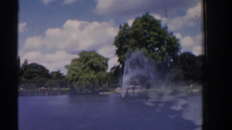 1961 Ireland 8mm frame with a distant pond fountain, tree line, blue sky, and visible film wear along the frame edge.