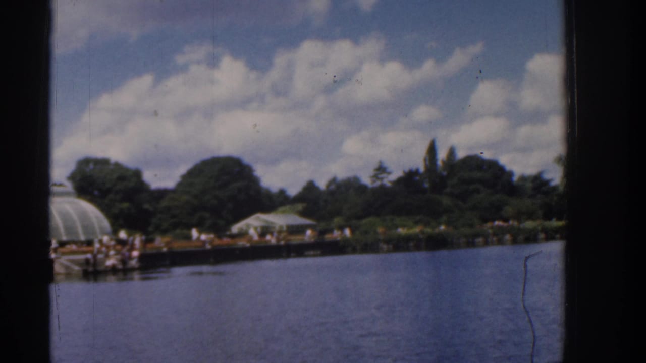 1961 Ireland lakeside view with a glasshouse-like structure, white shelter, water, trees, and distant people along the promenade.