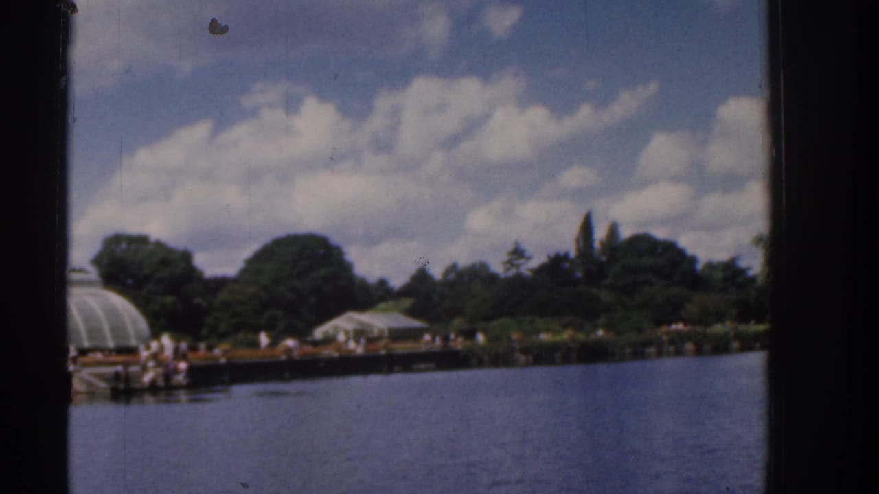 A wider 8mm frame of the Irish lakeside promenade, showing water, public garden structures, trees, clouds, and small distant visitor figures.
