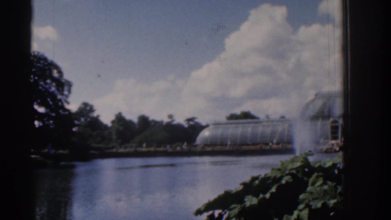 1961 Ireland 8mm frame of a lake garden view through a dark edge, with a glass conservatory, fountain spray, trees, and bright clouds.