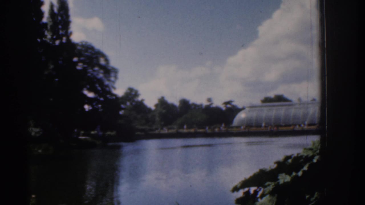 1961 Ireland garden lake view with a glass conservatory across the water, dark trees, distant visitors, and large white clouds.