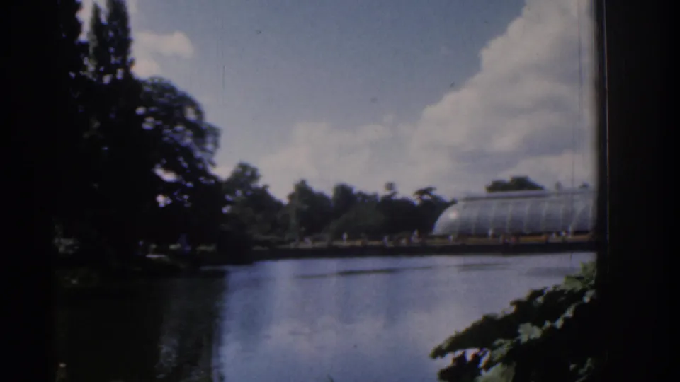 1961 Ireland garden lake view with a glass conservatory across the water, dark trees, distant visitors, and large white clouds.