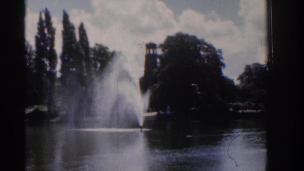 1961 Ireland 8mm frame of a lake fountain spraying in front of tall trees and a tower-like garden structure, with clouds overhead.