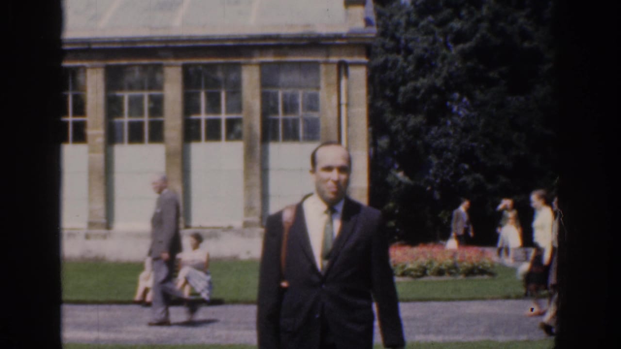 Suited man nears the camera while pedestrians move through a landscaped park behind him.