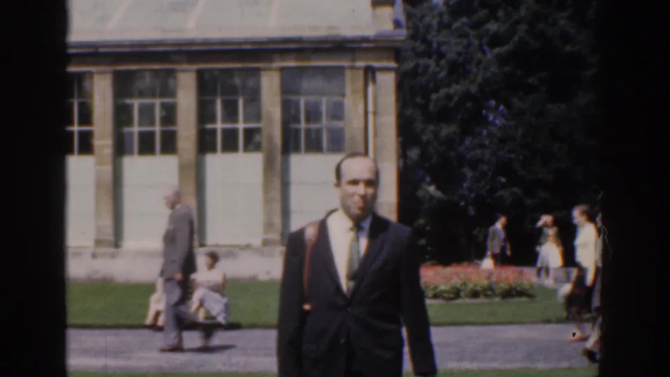 The suited man continues walking toward the camera in Ireland, with pedestrians, benches, flower beds, and trees behind him.