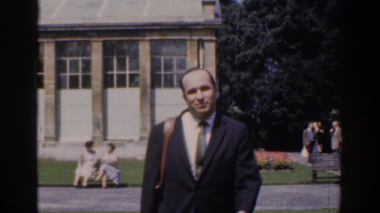 A smiling suited man with a shoulder bag approaches in the 1961 Ireland park scene, with visitors and building windows behind him.
