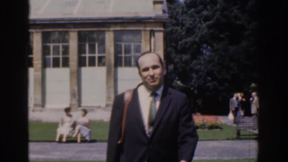 A smiling suited man with a shoulder bag approaches in the 1961 Ireland park scene, with visitors and building windows behind him.