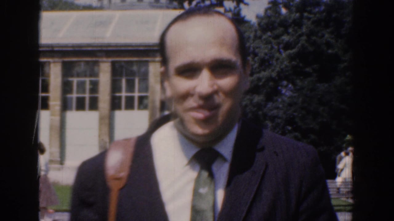 In 1961 Ireland home-movie footage, a suited man with a shoulder strap faces the camera in a landscaped park setting.