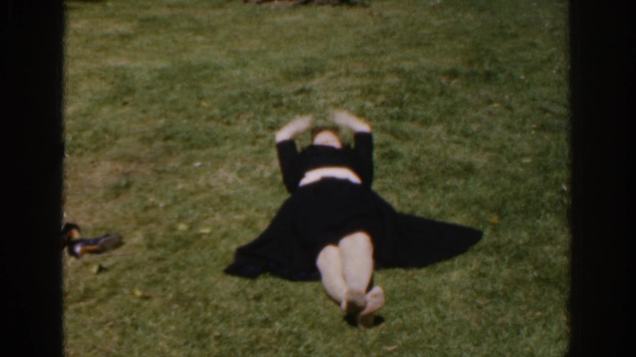 The woman lies on a grassy yard in 1961 Ireland footage, stretching her arms above her head.