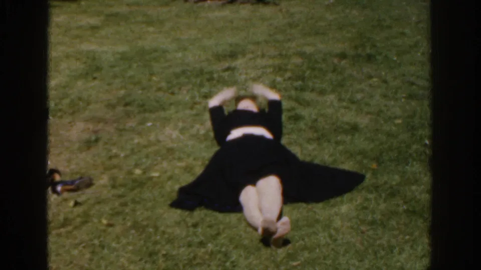 The woman lies on a grassy yard in 1961 Ireland footage, stretching her arms above her head.