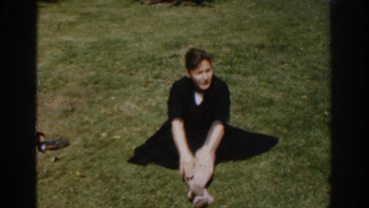 In the home-movie frame, the woman sits forward on grass and reaches toward her feet during a stretch.