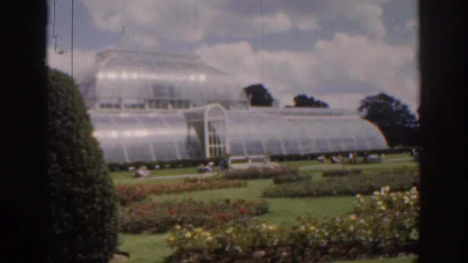 1961 Ireland 8mm frame showing a glass greenhouse with an arched doorway, formal garden beds, trees, and tiny visitors in the grounds.