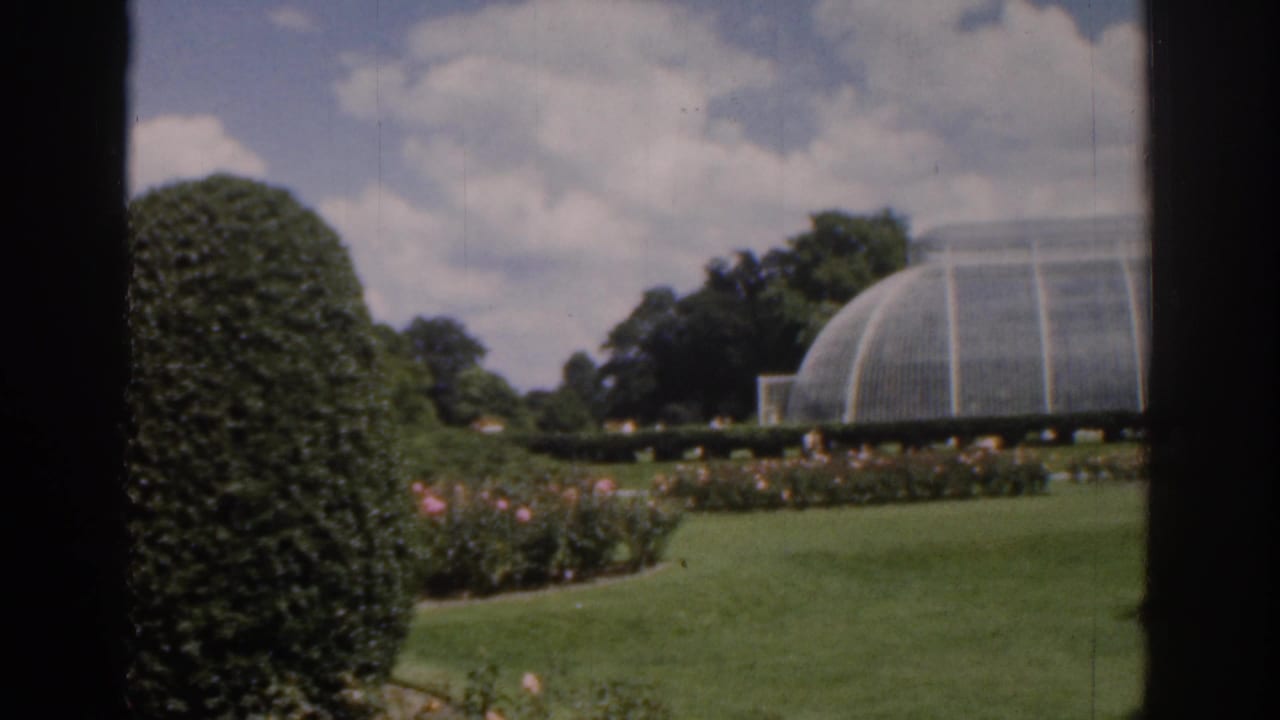 Large glass greenhouse behind a lawn, flower beds, trees, and a rounded hedge.