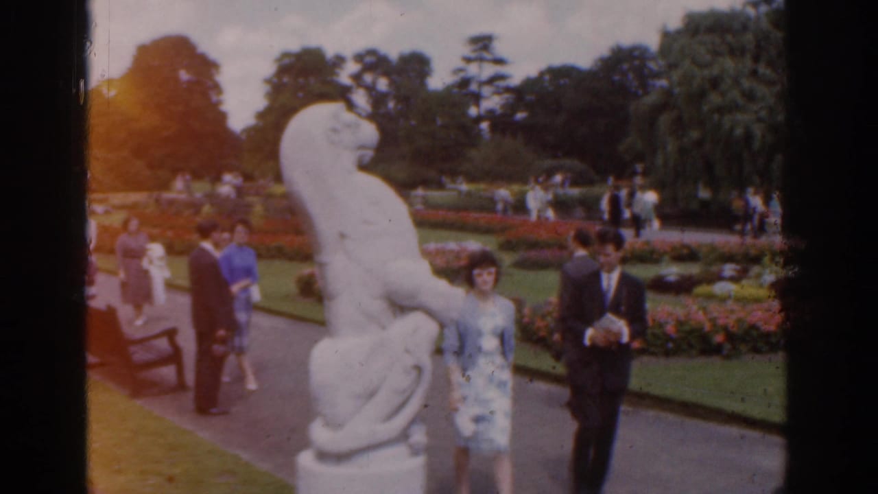 1961 Ireland home-movie frame showing visitors near a white animal sculpture, flowerbeds, paths, and mature trees.
