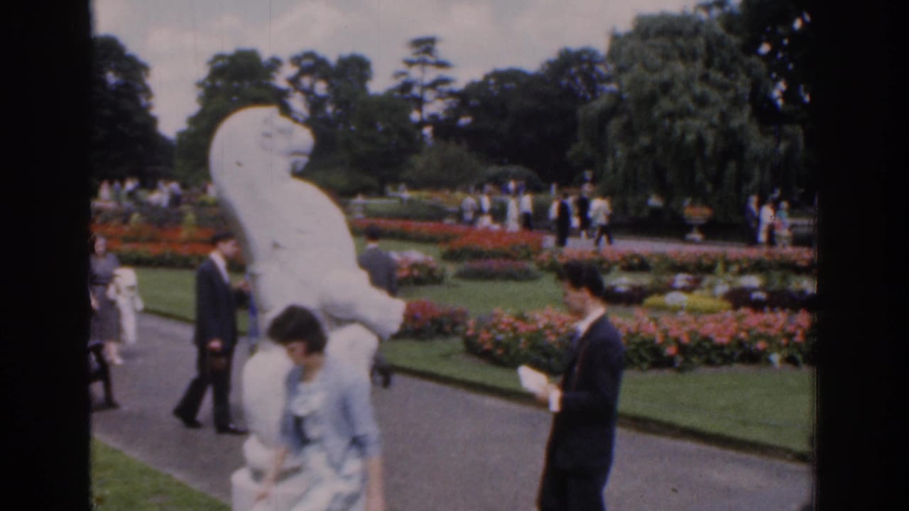 A suited man and woman stand beside a white sculpture in a flower garden.