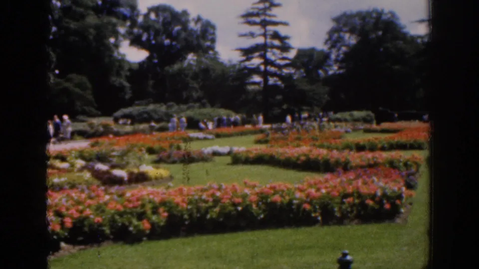 1961 Ireland garden frame showing colorful flowerbeds, lawns, paths, trees, and small groups of distant visitors.
