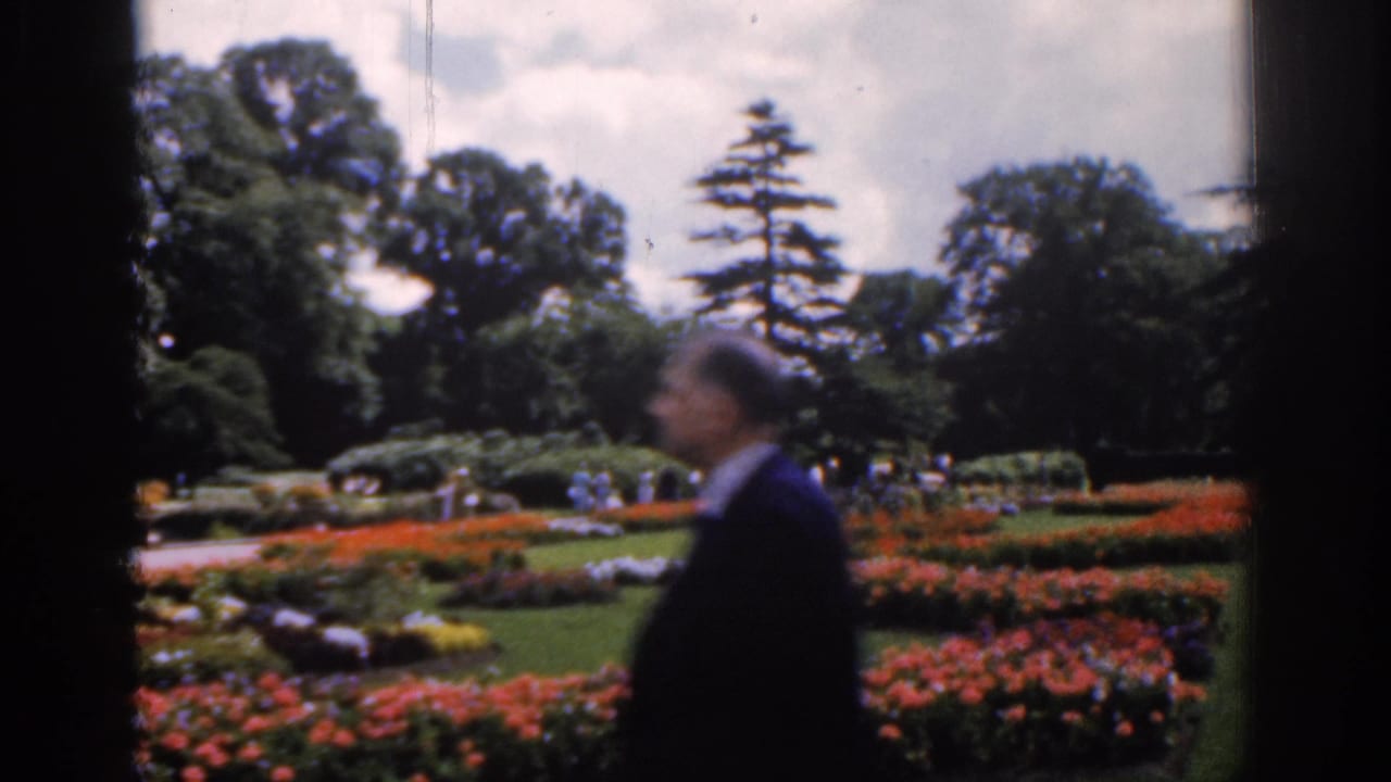 Man walking past flower beds in a formal garden with visitors and trees behind him.
