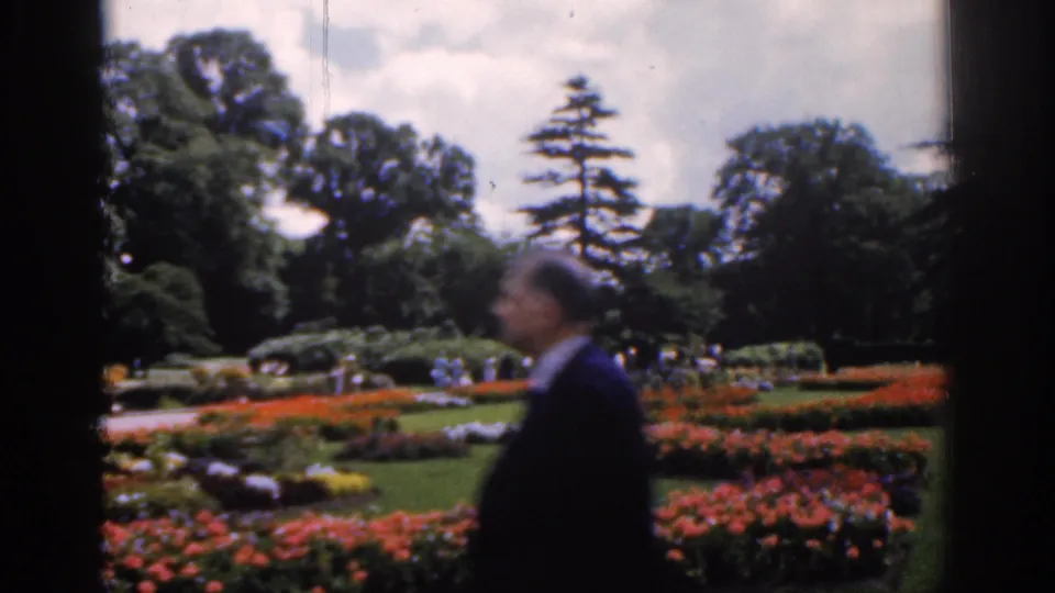 A man passes in front of flower beds in a 1961 Ireland garden scene, with lawns, paths, visitors, and mature trees in the background.