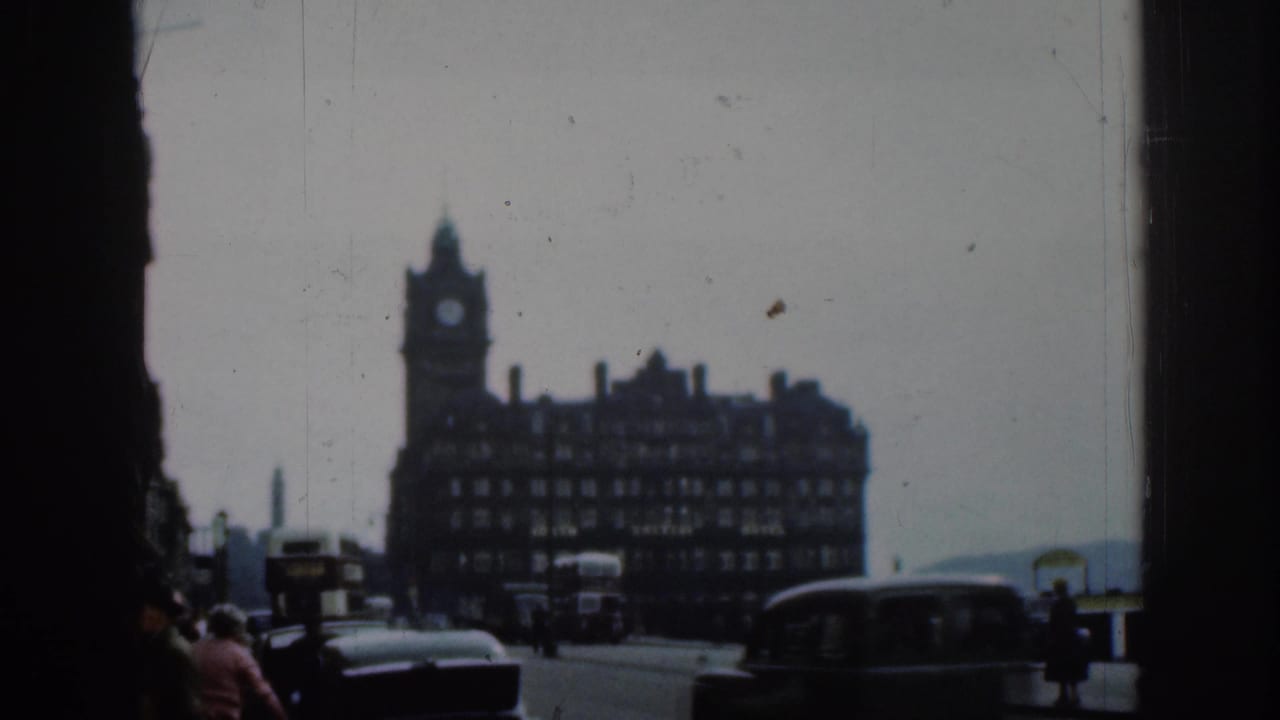 Clock-tower hotel facade seen across a London street with cars, buses, pedestrians, and dark framing at the sides.