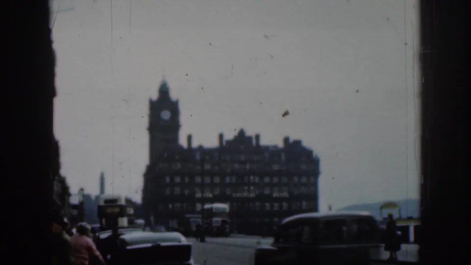 1961 London 8mm frame showing a clock-tower hotel facade across a street, with vintage vehicles and pedestrians visible through a dark foreground frame.