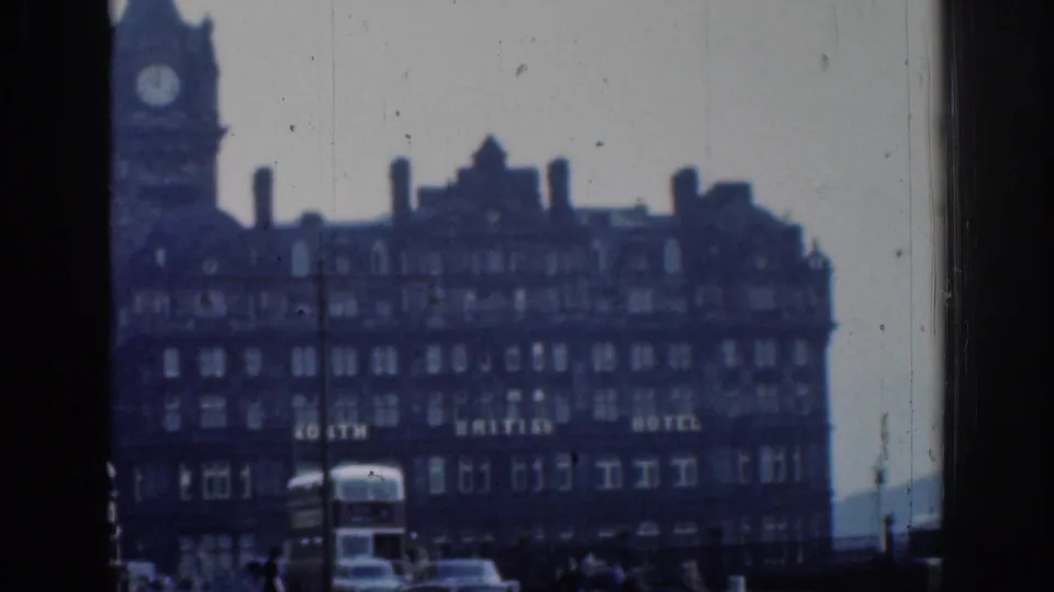 1961 London 8mm frame showing the hotel facade and clock tower with partial lettering, buses and cars below, and dark foreground framing.