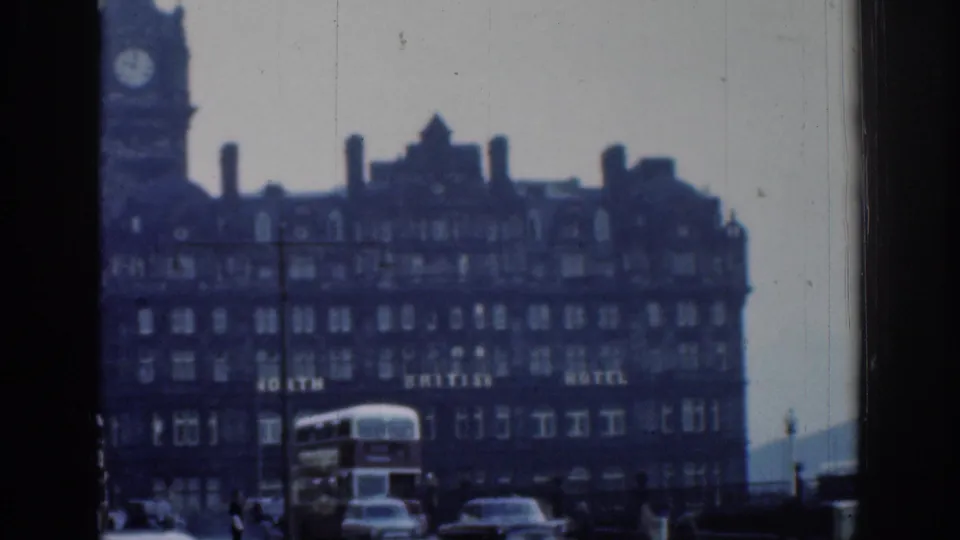 1961 London street view with a large hotel facade, clock tower, cars, and a double-decker bus seen through soft 8mm exposure.