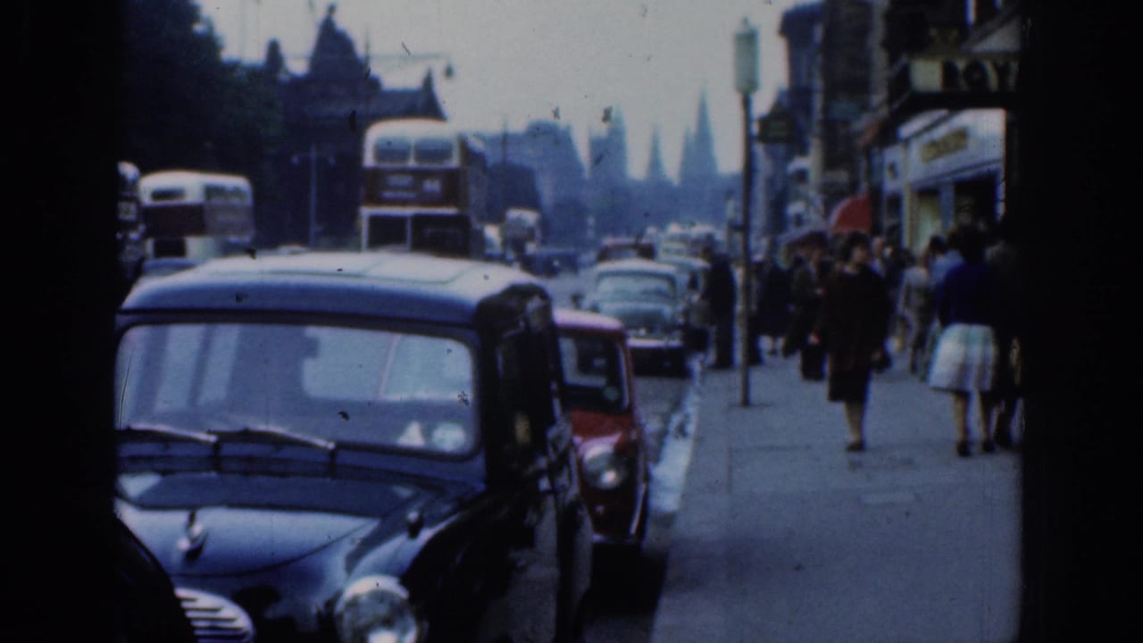 1961 London street scene with a black compact car in front, double-decker buses, storefronts, and pedestrians along the pavement.