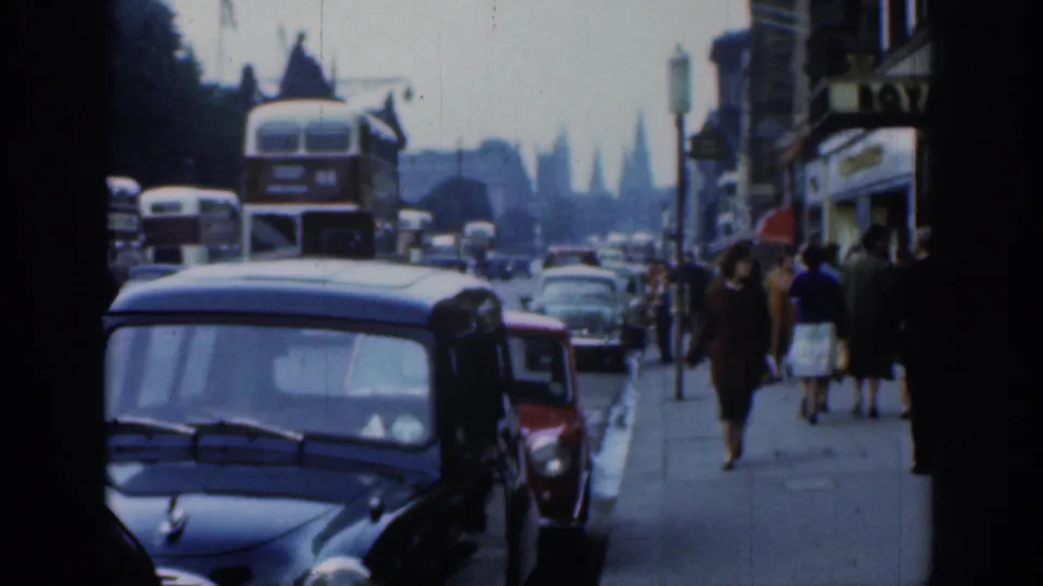 Street-level 1961 London view with a black compact car, red car, double-decker buses, pedestrians, and commercial shopfront signs.