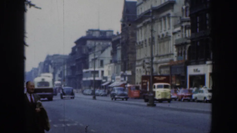 1961 London street view on 8mm film, showing classic traffic, storefront buildings, and a suited pedestrian near the camera.