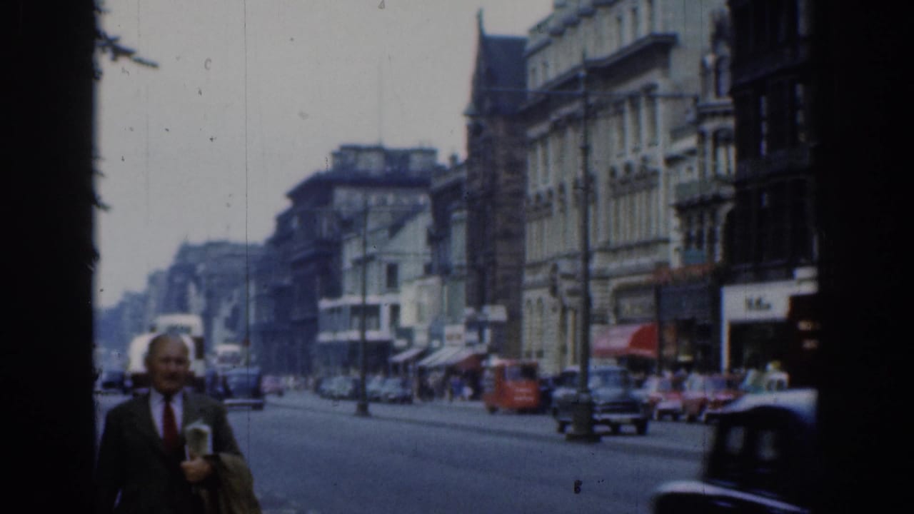 1961 London street scene with vintage cars, pedestrians near shops, and a suited man walking close to the camera.