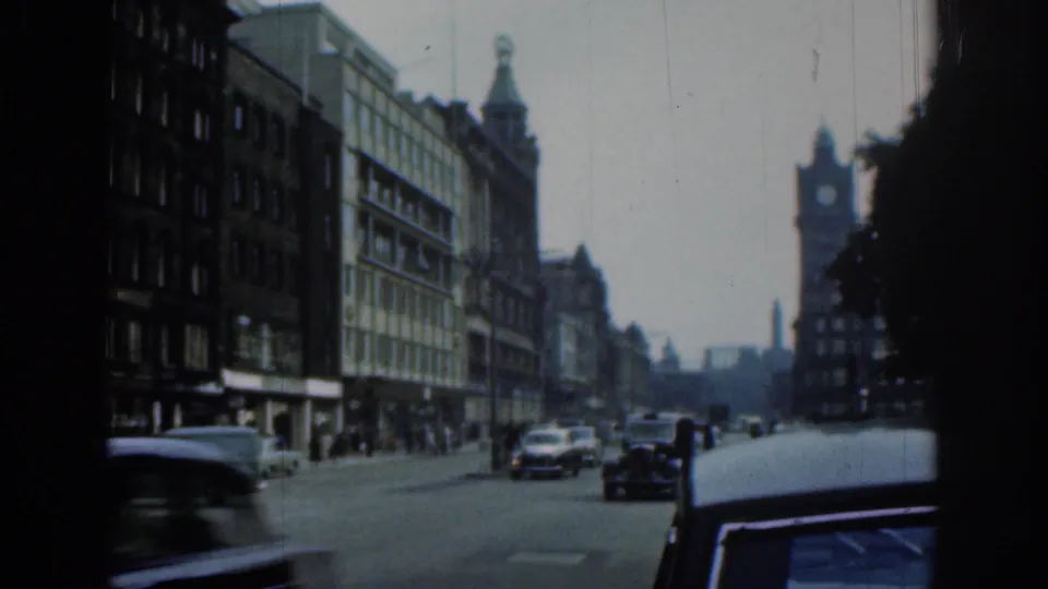 1961 London roadway seen from the curb, with classic cars, pedestrians, tall commercial buildings, and a clock tower in the distance.