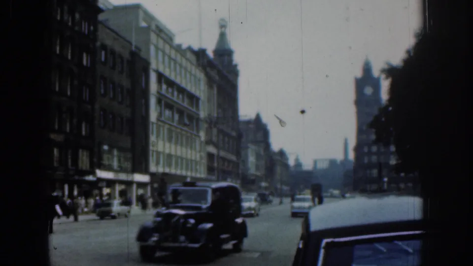 1961 London street view with a black taxi in the roadway, other period cars, pedestrians, and a distant clock tower between buildings.