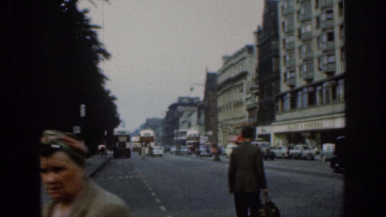 A passerby crosses the foreground of a 1961 London street scene with cars, buses, buildings, and a shaded sidewalk behind.