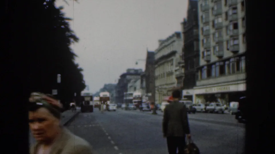 A passerby crosses the foreground of a 1961 London street scene with cars, buses, buildings, and a shaded sidewalk behind.