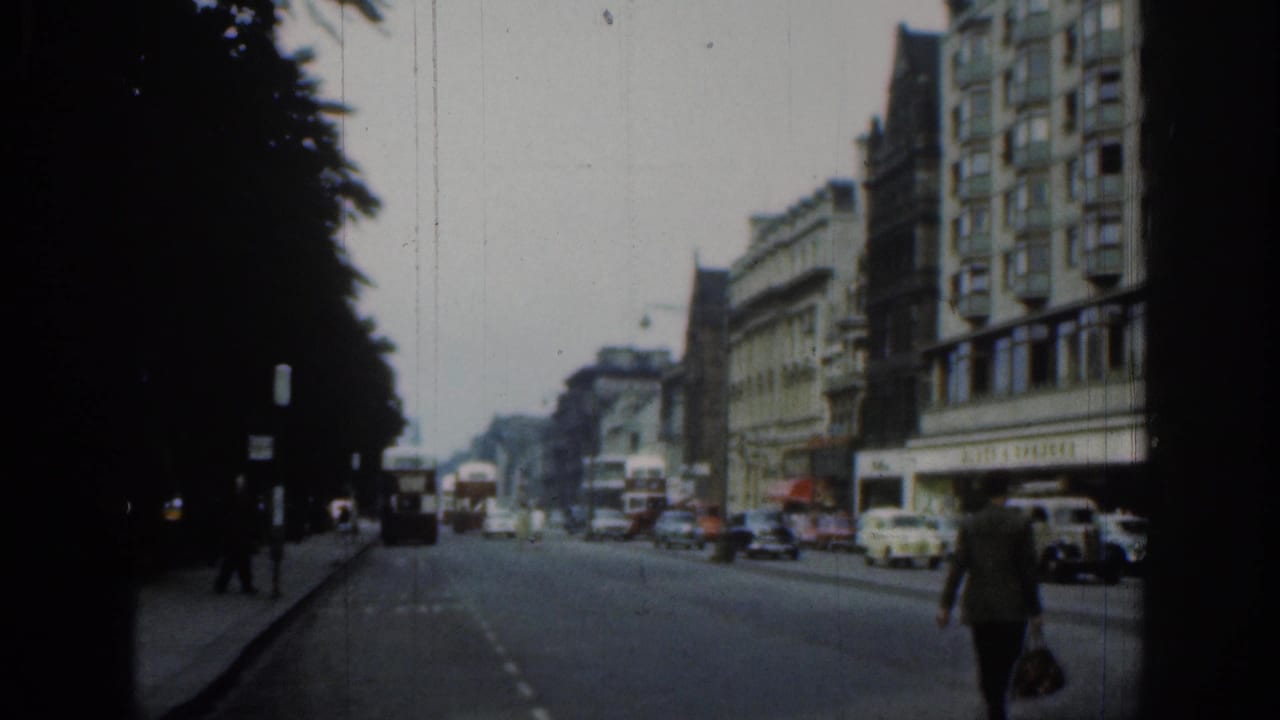 1961 London road scene with period cars, buses, pedestrians on the pavement, dark trees, and multi-story commercial buildings.
