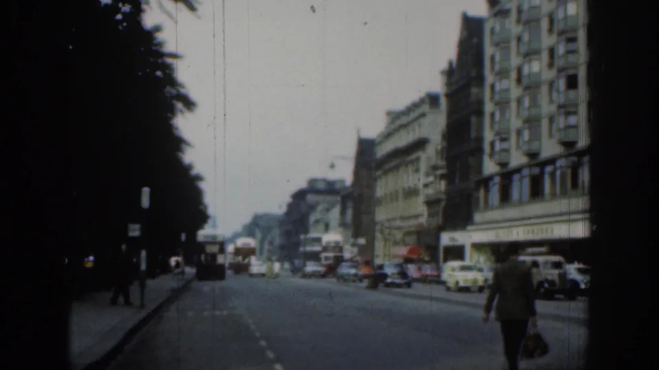 1961 London road scene with period cars, buses, pedestrians on the pavement, dark trees, and multi-story commercial buildings.