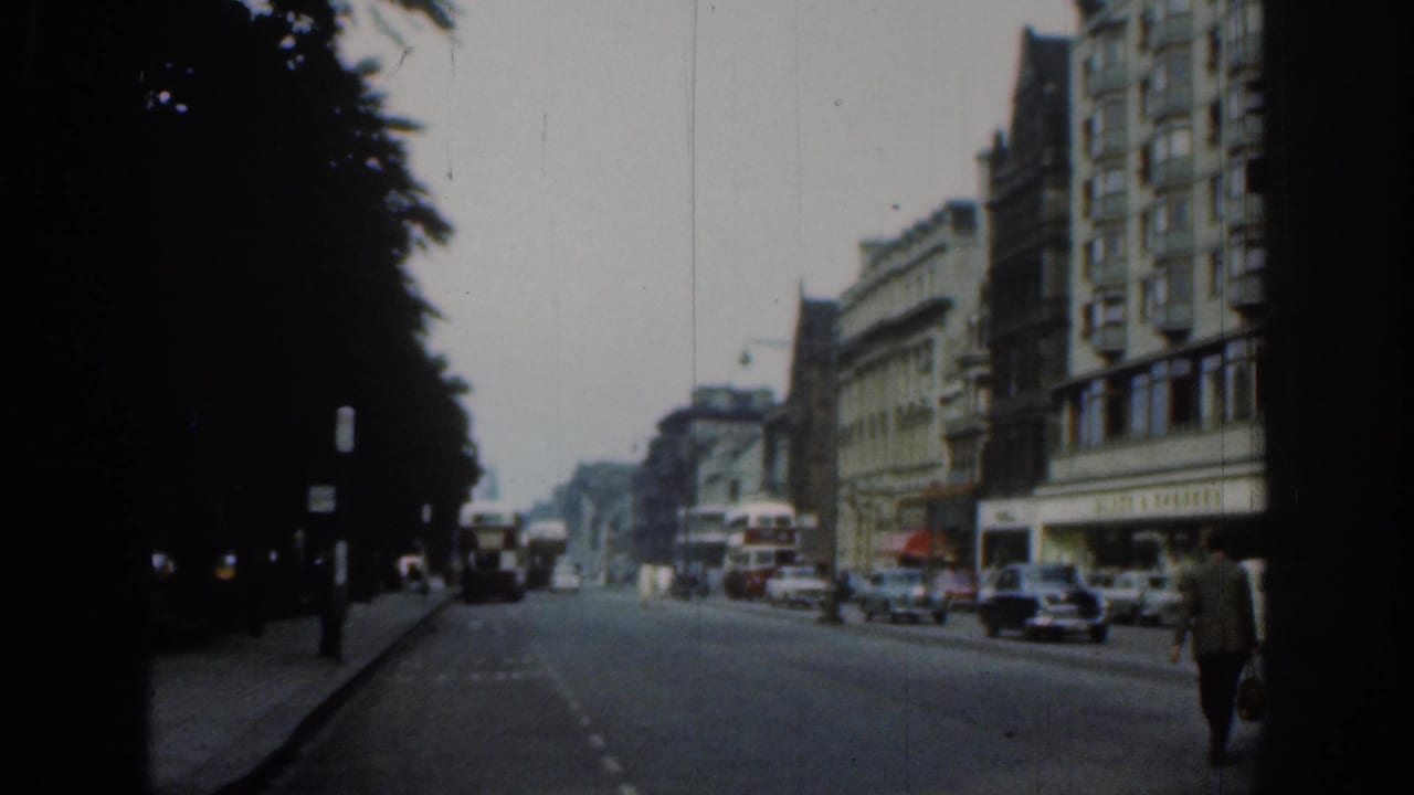 Final 1961 London street frame showing cars, buses, shopfronts, a dark tree line, and a pedestrian walking near the right edge.